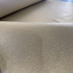 May include: A roll of shimmering gold fabric with a textured surface. The fabric is partially unrolled, revealing a lighter, cream-colored backing. The material has a subtle sparkle, catching the light.