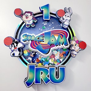 May include: A Space Jam themed cake topper with a blue, white, and red color scheme. The topper features the characters Bugs Bunny, Tweety Bird, and Taz holding basketballs. The topper also includes the words "Space Jam" and the number "1".