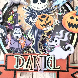 Puede incluir: Un adorno para pastel con personajes de Pesadilla Antes de Navidad. El adorno incluye a Jack Skellington, Sally, Zero, Oogie Boogie y una calabaza. El adorno est&aacute; decorado con purpurina y el nombre "DANIEL" est&aacute; escrito en la parte inferior.