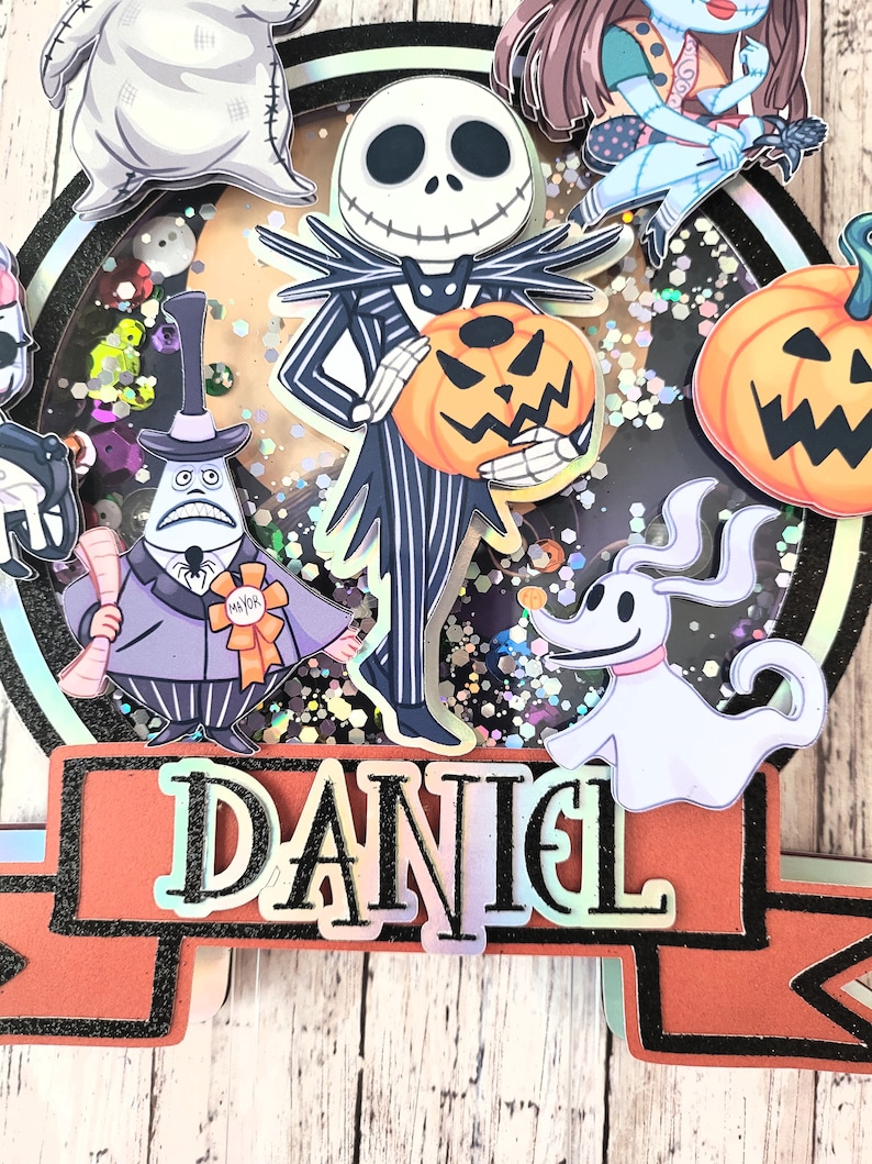 Puede incluir: Un adorno para pastel con personajes de Pesadilla Antes de Navidad. El adorno incluye a Jack Skellington, Sally, Zero, Oogie Boogie y una calabaza. El adorno est&aacute; decorado con purpurina y el nombre "DANIEL" est&aacute; escrito en la parte inferior.