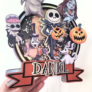 Puede incluir: Un adorno para pastel de varios niveles con personajes de la pel&iacute;cula Pesadilla antes de Navidad. El adorno incluye a Jack Skellington, Sally, Oogie Boogie, Zero y el alcalde. El adorno est&aacute; decorado con brillantina y una pancarta que dice "DANIEL".