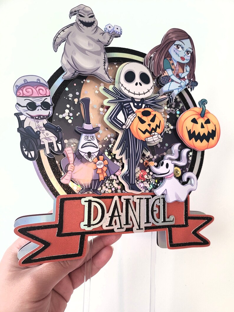 Puede incluir: Un adorno para pastel de varios niveles con personajes de la pel&iacute;cula Pesadilla antes de Navidad. El adorno incluye a Jack Skellington, Sally, Oogie Boogie, Zero y el alcalde. El adorno est&aacute; decorado con brillantina y una pancarta que dice "DANIEL".