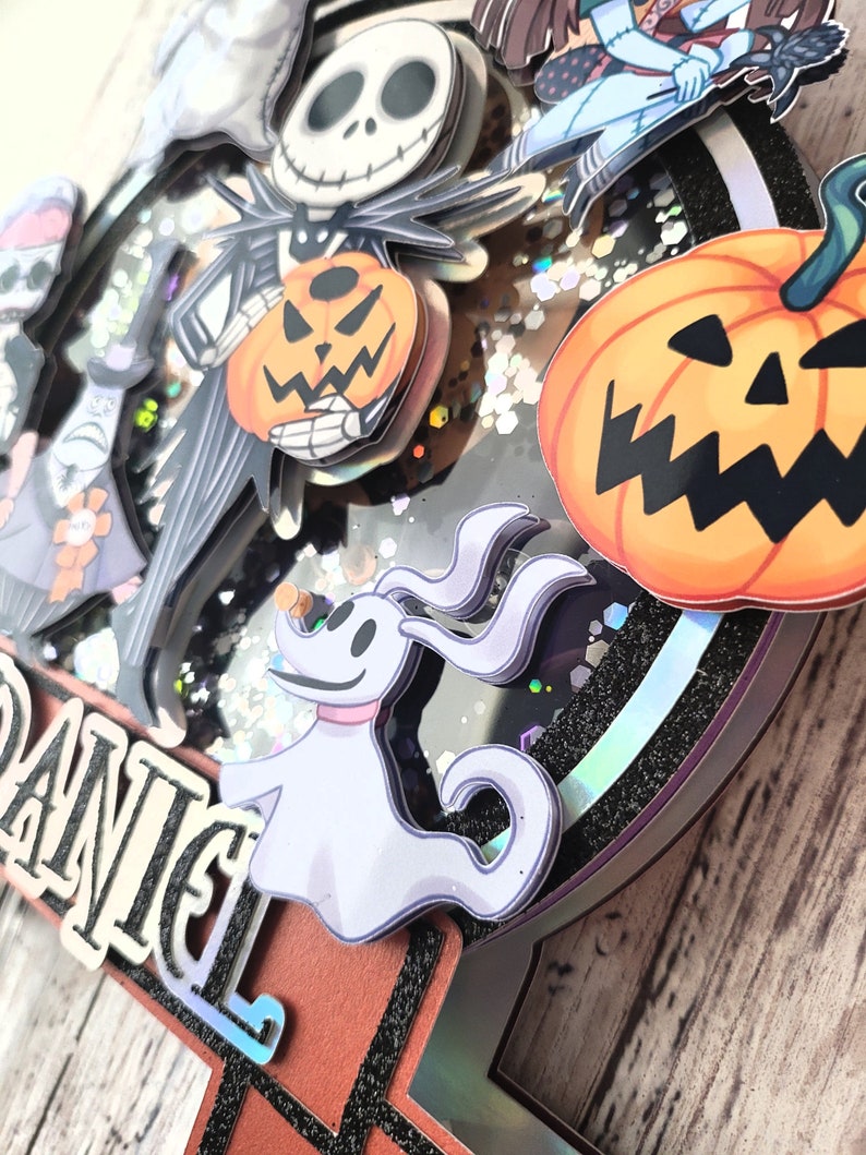 Puede incluir: Un adorno para pastel en blanco y negro con personajes de la pel&iacute;cula Pesadilla antes de Navidad. El adorno incluye a Jack Skellington, Sally, Zero el perro fantasma y una calabaza. El adorno est&aacute; decorado con purpurina y tiene el nombre "Daniel" escrito en &eacute;l.