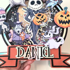 Puede incluir: Un adorno redondo para pastel con un fondo brillante que presenta personajes de Pesadilla Antes de Navidad. Los personajes incluyen a Jack Skellington, Sally, Zero, Oogie Boogie y el alcalde. El adorno tambi&eacute;n incluye una pancarta con el nombre "DANIEL".
