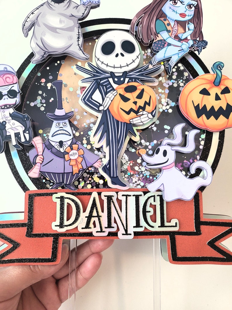 Puede incluir: Un adorno redondo para pastel con un fondo brillante que presenta personajes de Pesadilla Antes de Navidad. Los personajes incluyen a Jack Skellington, Sally, Zero, Oogie Boogie y el alcalde. El adorno tambi&eacute;n incluye una pancarta con el nombre "DANIEL".