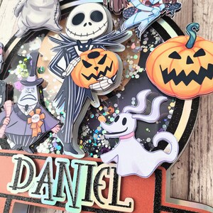 Puede incluir: Un adorno para pastel con tem&aacute;tica de Halloween con un personaje de rayas blancas y negras sosteniendo una linterna de calabaza, un fantasma blanco, un monstruo gris, una calabaza y una ni&ntilde;a con cabello casta&ntilde;o. El adorno est&aacute; decorado con brillantina y el nombre "DANIEL" est&aacute; escrito en una pancarta.