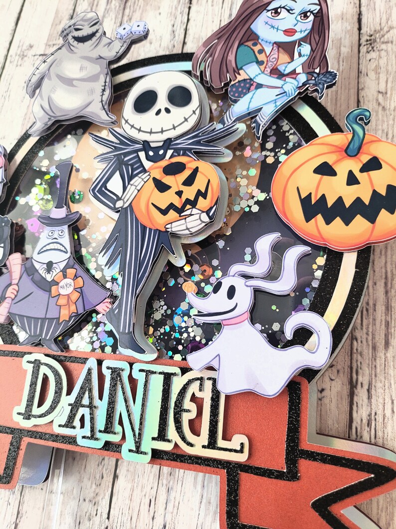 Puede incluir: Un adorno para pastel con tem&aacute;tica de Halloween con un personaje de rayas blancas y negras sosteniendo una linterna de calabaza, un fantasma blanco, un monstruo gris, una calabaza y una ni&ntilde;a con cabello casta&ntilde;o. El adorno est&aacute; decorado con brillantina y el nombre "DANIEL" est&aacute; escrito en una pancarta.