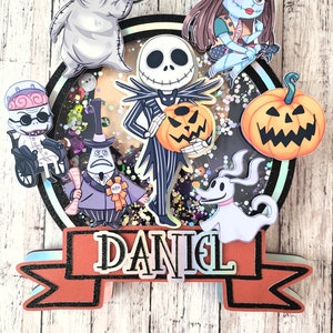 Puede incluir: Un adorno redondo para pastel con un fondo brillante que presenta personajes de Pesadilla Antes de Navidad. Los personajes incluyen a Jack Skellington, Sally, Zero, Oogie Boogie y el Rey Calabaza. El adorno tiene una pancarta con el nombre "DANIEL" en ella.