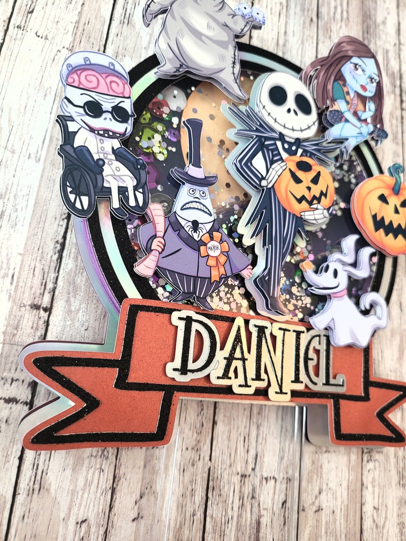 Puede incluir: Un adorno para pastel con personajes de la pel&iacute;cula Pesadilla antes de Navidad. El adorno incluye a Jack Skellington, Sally, Oogie Boogie, Zero y el alcalde. El adorno est&aacute; decorado con brillantina y tiene una pancarta que dice "DANIEL".