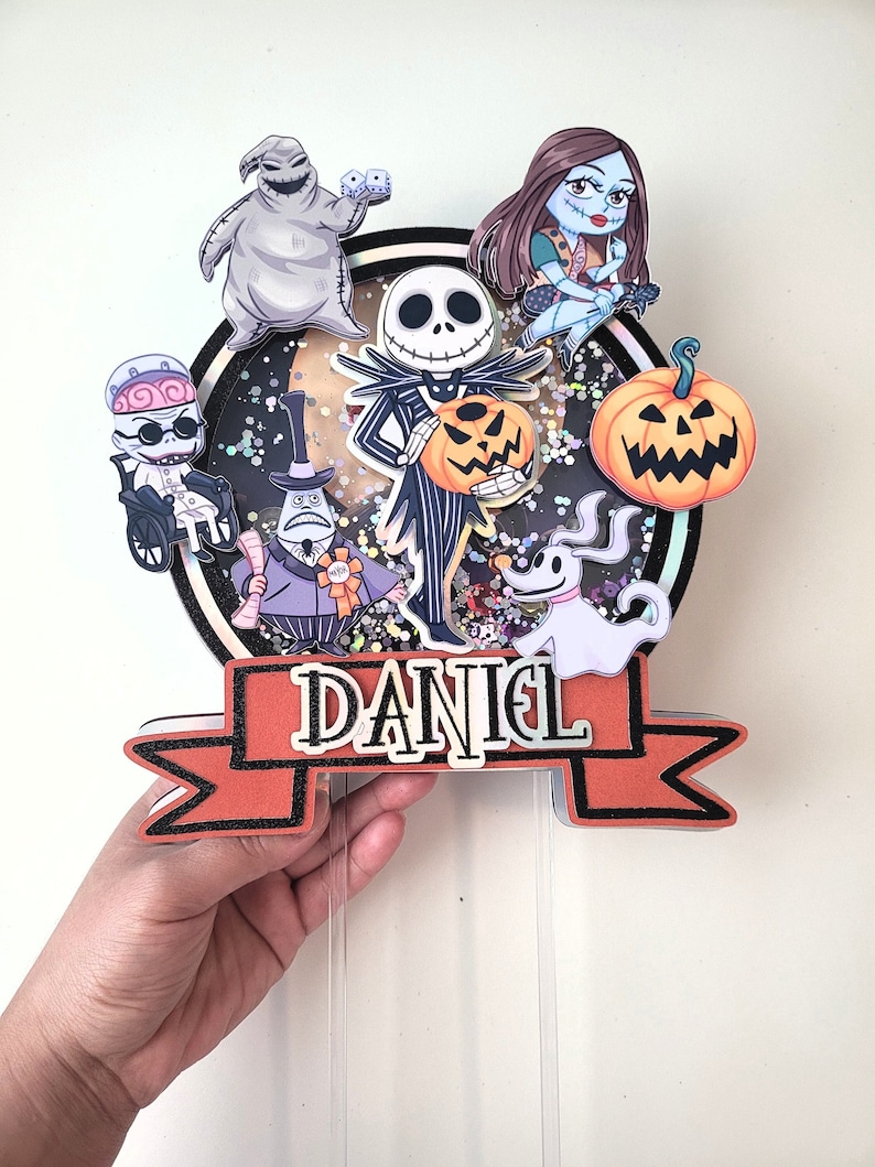 Puede incluir: Un adorno para pastel con personajes de la pel&iacute;cula Pesadilla antes de Navidad. El adorno incluye a Jack Skellington, Sally, Oogie Boogie, Zero y el Rey Calabaza. El adorno est&aacute; decorado con purpurina y tiene una pancarta que dice "DANIEL".