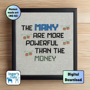 May include: A framed cross-stitch artwork featuring the phrase "THE MANY ARE MORE POWERFUL THAN THE MONEY." The word "MANY" is in blue. The frame is black. The art is on a light beige fabric. The image also includes the text "Human made art (NO AI)" and "Digital Download."