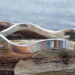May include: A polished silver bangle bracelet featuring a continuous wavy, undulating design. The metal surface is smooth and reflective, with a visible hinge mechanism on one side and a 925 hallmark stamped on the inner band of the jewelry piece.