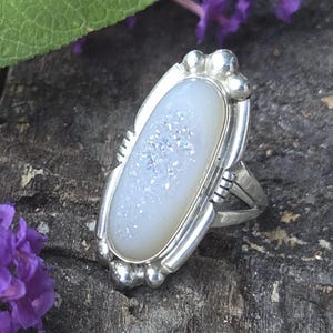May include: A sterling silver ring featuring an elongated oval druzy stone with a shimmering white surface. The metal setting includes decorative silver beads at the top and bottom, with grooved accents along the sides of the central stone.