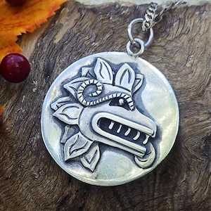 May include: A circular silver pendant featuring a relief carving of a stylized creature head with open jaws and leaf-like elements. The pendant is attached to a silver chain link and has a polished finish with darkened recessed areas.