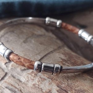 May include: A silver bangle bracelet with a brown, textured leather band. The bracelet has silver accents and a simple, elegant design. The leather band appears to be made of snakeskin.