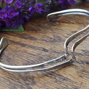 May include: A silver-toned metal cuff bracelet featuring a double-band design that curves into a wave-like shape. The bracelet has an open back and a smooth, polished finish, showcasing a continuous flowing structure that wraps around the wrist.
