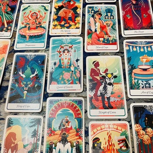 Pode incluir: Um conjunto de cartas de tar&ocirc; com ilustra&ccedil;&otilde;es coloridas que retratam v&aacute;rias cenas e personagens. As cartas apresentam uma variedade de cores, incluindo azul, verde, vermelho e dourado. As cartas est&atilde;o dispostas em um padr&atilde;o de grade em uma superf&iacute;cie com um padr&atilde;o azul e branco.
