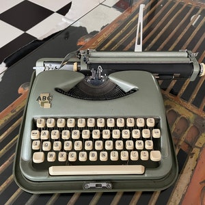 May include: A vintage, light green typewriter with white keys and the letters "ABC" on the front. The keyboard layout is AZERTY. The typewriter sits on a reflective surface with a black and white checkered pattern in the background.
