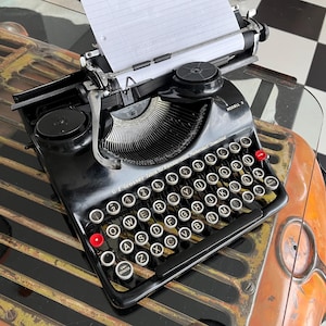 May include: A vintage black typewriter with white keys and red accents. The typewriter has a paper inserted and is sitting on a metal and glass surface. The keys are arranged in a QWERTY layout.
