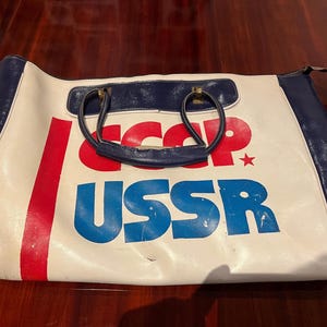 Op de afbeelding: Een witte reistas met blauwe en rode accenten. De tas heeft een blauwe handgreep en de tekst "USSR" gedrukt in blauwe letters.