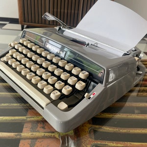 May include: A gray Brother typewriter with a white paper in the carriage. The typewriter has a black keyboard with white keys and a black and white design.