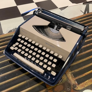 Puede incluir: Una máquina de escribir vintage Remington Elektro 111 azul y beige con una palanca de retorno de carro negra. La máquina de escribir está sobre una mesa de vidrio con una rejilla metálica debajo.