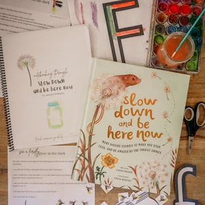 May include: An open book titled "Slow Down and Be Here Now" with a dandelion illustration, alongside a spiral notebook and educational materials. A watercolor palette, scissors, and the letter "F" are also visible. The theme is nature and learning.