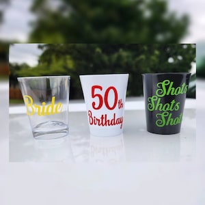 May include: Three clear plastic shot glasses with different designs. The first glass has the word "Bride" in yellow lettering. The second glass has the text "50th Birthday" in red lettering. The third glass has the text "Shots Shots Shot" in green lettering.