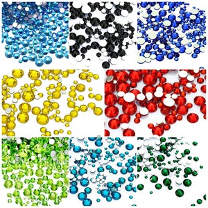 May include: Assortment of colorful rhinestones in various colors, including blue, black, yellow, red, green, and turquoise. The rhinestones are scattered on a white background.