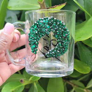 May include: A clear glass mug with a handle, featuring a black and white line drawing of a woman's face with green rhinestones outlining her hair.