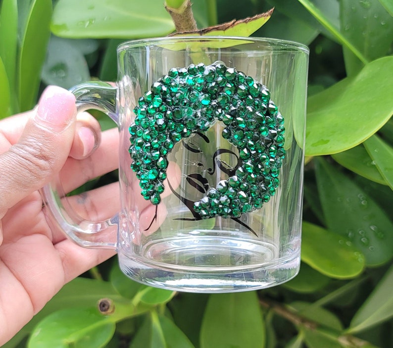 May include: A clear glass mug with a handle, featuring a black and white line drawing of a woman's face with green rhinestones outlining her hair.