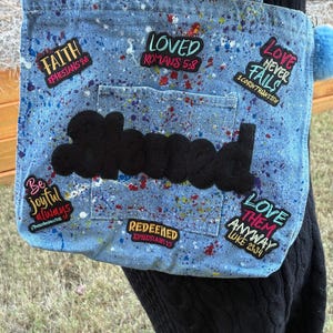 May include: A light blue denim tote bag with colorful paint splatters and patches with inspirational quotes. The bag features a black cloud-shaped applique and a blue pom-pom. The patches include phrases like "Faith" and "Loved".