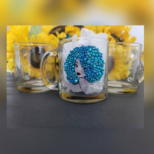 May include: A clear glass mug with a blue rhinestone design of a woman's face with an afro. The mug has a handle and is sitting on a white surface.