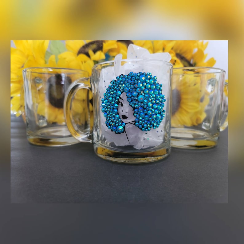 May include: A clear glass mug with a blue rhinestone design of a woman's face with an afro. The mug has a handle and is sitting on a white surface.