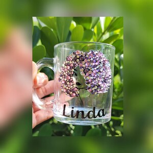 May include: A clear glass mug with a black silhouette of a woman with a pink and purple rhinestone afro. The mug has the name "Linda" written in black lettering on the bottom.