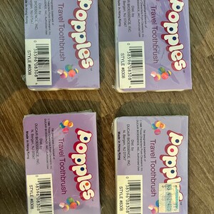 Puede incluir: Cuatro cepillos de dientes de viaje de color morado y blanco con el logotipo de Popples y el texto "Travel Toothbrush". El empaque presenta una imagen de dibujos animados de los personajes de Popples.