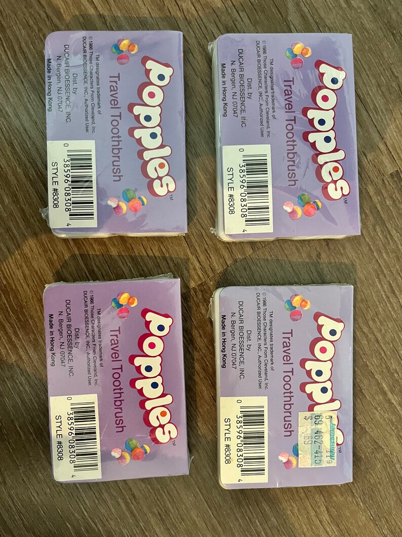 Puede incluir: Cuatro cepillos de dientes de viaje de color morado y blanco con el logotipo de Popples y el texto "Travel Toothbrush". El empaque presenta una imagen de dibujos animados de los personajes de Popples.