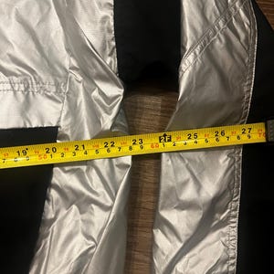 Może przedstawiać: Srebrno-czarna kurtka o wymiarach około 69 cm. Kurtka ma błyszczący, metaliczny wygląd i jest wykonana z lekkiego materiału. Kurtka ma czarne panele po bokach.