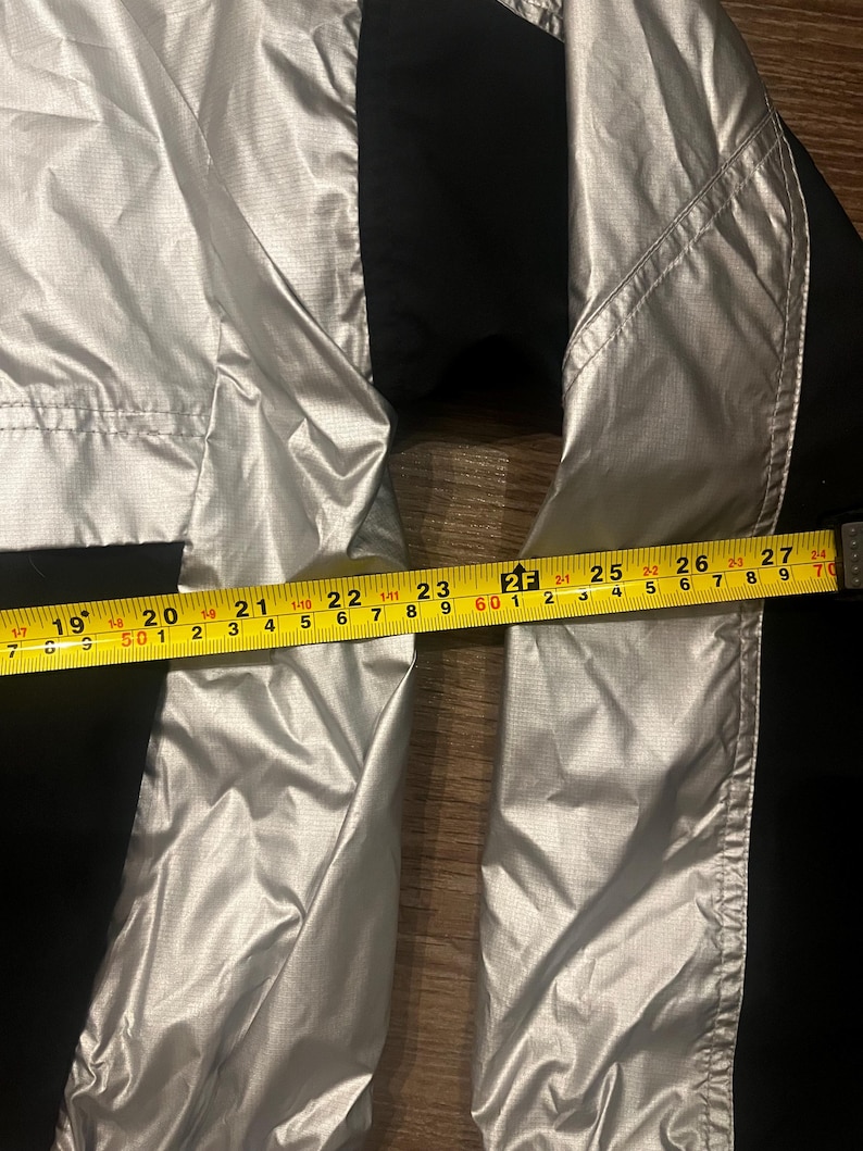 Może przedstawiać: Srebrno-czarna kurtka o wymiarach około 69 cm. Kurtka ma błyszczący, metaliczny wygląd i jest wykonana z lekkiego materiału. Kurtka ma czarne panele po bokach.