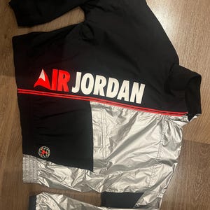Może przedstawiać: Czarna i srebrna kurtka dresowa Air Jordan. Kurtka posiada napis "AIR JORDAN" w kolorze białym z czerwoną linią akcentującą. Czerwono-czarne logo globu znajduje się na lewym rękawie. Kurtka wykonana jest z błyszczącego, wodoodpornego materiału.
