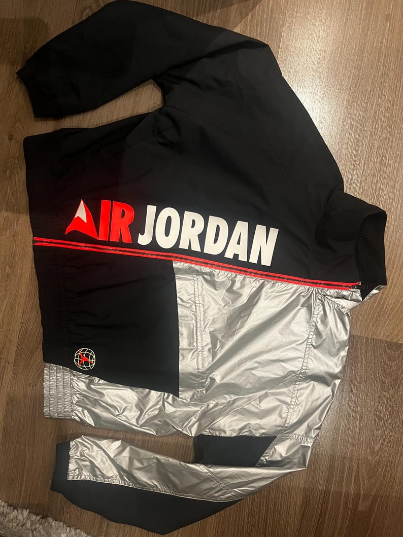 Może przedstawiać: Czarna i srebrna kurtka dresowa Air Jordan. Kurtka posiada napis "AIR JORDAN" w kolorze białym z czerwoną linią akcentującą. Czerwono-czarne logo globu znajduje się na lewym rękawie. Kurtka wykonana jest z błyszczącego, wodoodpornego materiału.