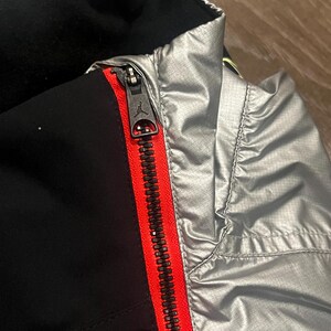 Może przedstawiać: Zbliżenie czarnej i srebrnej kurtki z czerwonym zamkiem błyskawicznym. Zamek jest zamknięty i posiada czarny suwak z logo Jumpman. Kurtka ma czarne wnętrze i srebrny wygląd zewnętrzny.