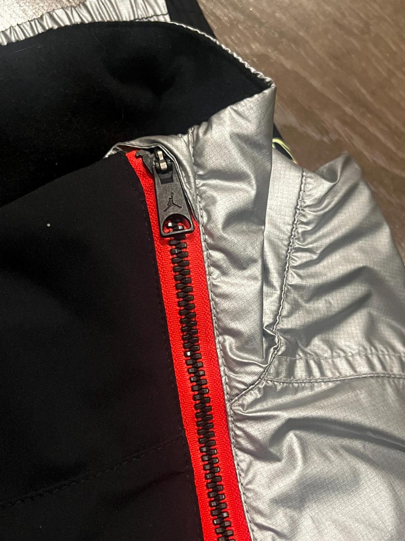 Może przedstawiać: Zbliżenie czarnej i srebrnej kurtki z czerwonym zamkiem błyskawicznym. Zamek jest zamknięty i posiada czarny suwak z logo Jumpman. Kurtka ma czarne wnętrze i srebrny wygląd zewnętrzny.