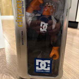 May include: A silver tin box with a clear plastic window showing a fully articulated action figure of a person wearing a black and blue jacket with a DC logo. The figure has orange hands and is standing on a black and white DC logo. The box has the text "citizen urban icon" and "DCSHOECOUSA" on it.