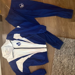 May include: A blue and white track suit, featuring a jacket and matching pants. The jacket has a white center panel with blue accents and a full zipper. The pants are solid blue. Both pieces display a team logo.
