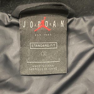 Może przedstawiać: Czarna metka odzieżowa z napisem "JORDAN" białymi wielkimi literami, czerwonym logo Jumpman i napisem "EST. 1985". Na metce widnieje r&oacute;wnież napis "STANDARD FIT" i "MADE IN CHINA / FABRIQUE EN CHINE".