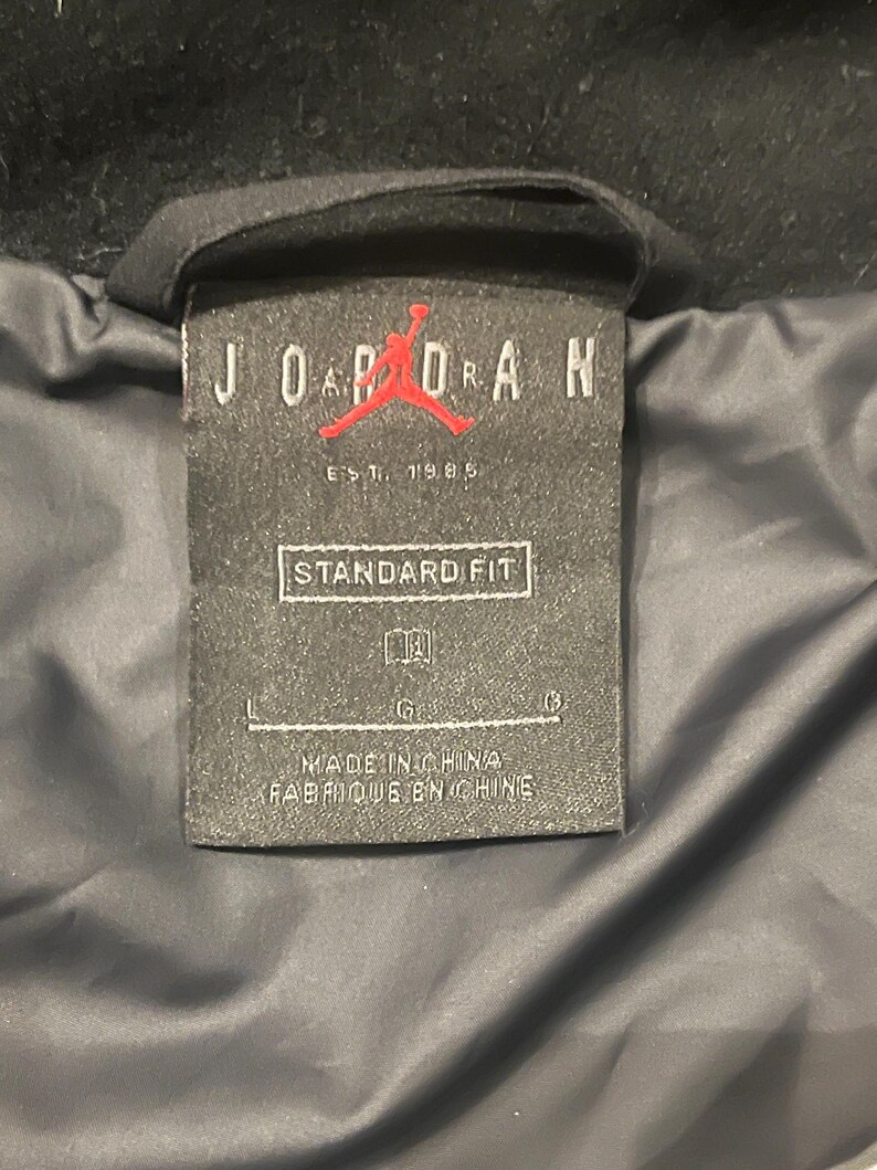 Może przedstawiać: Czarna metka odzieżowa z napisem "JORDAN" białymi wielkimi literami, czerwonym logo Jumpman i napisem "EST. 1985". Na metce widnieje r&oacute;wnież napis "STANDARD FIT" i "MADE IN CHINA / FABRIQUE EN CHINE".