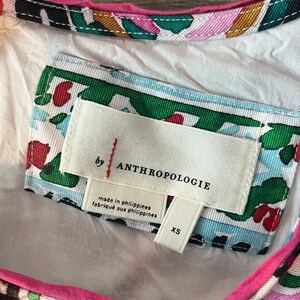 Może przedstawiać: Biała metka z czarnym tekstem, kt&oacute;ry brzmi "by Anthropologie" i "made in Philippines, fabrique aux Philippines". Metka ma r&oacute;wnież etykietę rozmiaru z napisem "XS". Metka jest przymocowana do białej tkaniny z kolorowym wzorem kwiatowym.