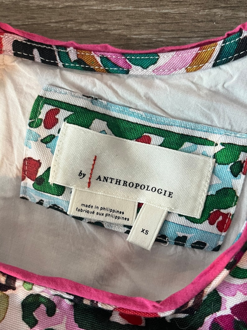 Może przedstawiać: Biała metka z czarnym tekstem, kt&oacute;ry brzmi "by Anthropologie" i "made in Philippines, fabrique aux Philippines". Metka ma r&oacute;wnież etykietę rozmiaru z napisem "XS". Metka jest przymocowana do białej tkaniny z kolorowym wzorem kwiatowym.