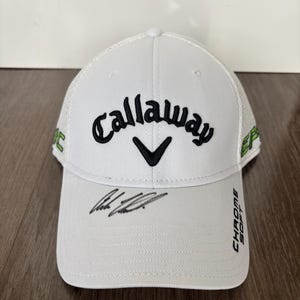 May include: White Callaway golf hat with a black logo and a black signature. The hat has a green and white mesh back and the text "Chrome Soft" on the side.